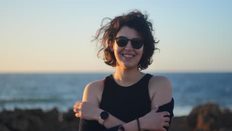 Young-woman-wearing-black-shades-smiling-at-the-camera,-ocean-behind-her-at-sunset