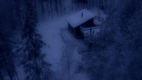 Cozy-small-cabin-at-night-in-snow-covered-forest,-aerial-drone-view