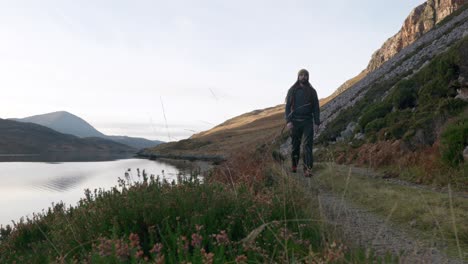 A-man-is-hiking-towards-the-camera-in-the-Scottish-Highlands,-with-a-cliff-and-a-sea-loch-in-the-background-while-heather-and-grass-gently-blows-in-the-wind-in-the-foreground