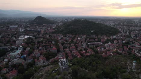 Plovdiv-cityscape-with-hills-covered-in-green,-sunset,-aerial-drone-view