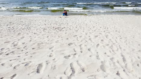 A-Mother-And-Child-Sitting-On-Beautiful-White-Sandy-Shore-Of-Baltic-Sea-With-Waves-Splashing-On-A-Sunny-Day