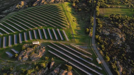 Photovoltaic-solar-power-station-on-rocky-hill-and-green-grass,-aerial,-tilt-down