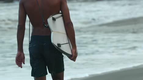 Man-walking-along-the-shore-of-the-beach-with-his-surfboard-in-hand,-medium-shot-in-slow-motion