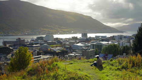 Person-Sitting-On-The-Grass-On-Sunny-Autumn-Morning-With-Scenery-Of-Townscape,-Tromsoysundet-And-Mountain-In-Tromso,-Norway
