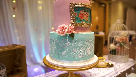 roses-designed-blue-and-pink-birthday-cake-on-Asian-event-close-up-moving-round-motion-on-a-stand-in-the-middle-of-the-room-with-fairy-lights-in-the-background-and-golden-accessories-laying-on-table