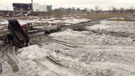 Aerial-shot-flying-above-a-dump-site-where-trucks-are-dropping-loads-of-snow-cleared-after-a-snowstorm,-while-bulldozers-plough-through-the-snow-spreading-it-across-a-field-in-Buffalo,-New-York