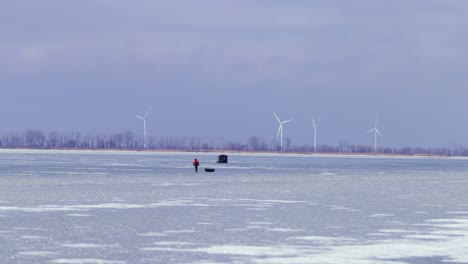 Fisher-men-waking-on-frozen-lake,-pulling-ice-sledge,-ice-fishing,-winter-cold-outdoor,-fishing-tent-on-frozen-lake,-mid-shot-Canada-wide-view-of-landscape,-windmill,-renewable-energy