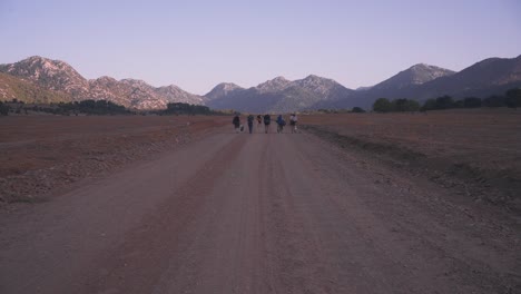 Establisher-wide-view-of-group-of-trekkers-with-backpacks-in-middle-of-vast-rural-road,-mountain-range-background-landscape