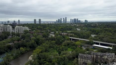 Drohne-Fliegt-über-Grünflächen,-Durch-Die-Ein-Fluss-In-Der-Innenstadt-Von-Toronto-Fließt,-Mit-Einer-Autobahn-In-Der-Ferne