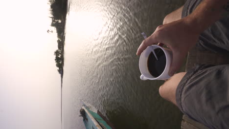 Vertikale-Ansicht-Eines-Jungen-Kaukasischen-Männlichen-Entdeckers,-Der-Bei-Sonnenuntergang-Eine-Tasse-Schwarzen-Kaffee-Trinkt,-Während-Er-Am-Flussufer-In-Senegal,-Afrika,-Sitzt
