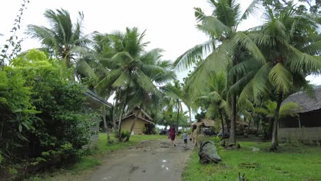 A-dynamic-shot-of-a-walking-family-towards-the-native-hut-surrounded-by-palm-trees-while-crossing-the-muddy-path