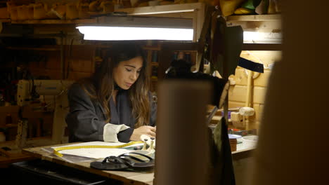 Female-shoemaker-working-at-workshop-desk-designing-stylish-footwear-under-spotlight-lamp