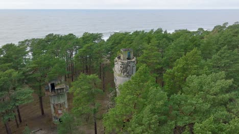 Establishing-aerial-view-of-old-Soviet-military-concrete-observation-watchtower,-pine-tree-forest,-Liepaja-,-military-heritage,-Nordic-woodland,-sea,-wide-drone-shot-moving-forward,-tilt-down