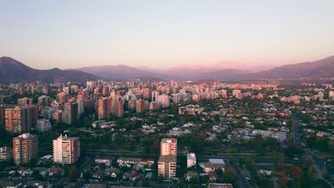 Aerial-orbit-of-Las-Condes,-a-wealthy-and-quiet-neighborhood-of-the-city,-illuminated-Andes-mountain-range-in-the-background,-epic-sunset