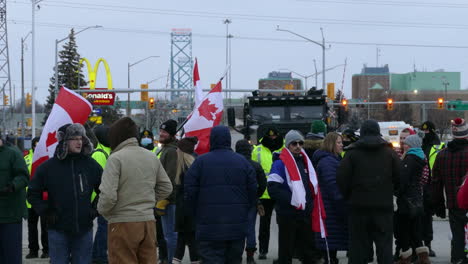 Toma-Estática-De-Manifestantes-Con-El-Convoy-De-La-Libertad-Ondeando-Banderas-Canadienses-Que-Protestan-Contra-El-Mandato-De-La-Vacuna-Covid-Y-El-Pasaporte-Con-El-Puente-Del-Embajador-Y-La-Policía-En-El-Fondo