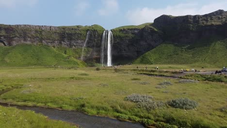 Flat-meadows-and-high-rocky-cliff-with-waterfall-in-Iceland,-low-altitude-aerial-view