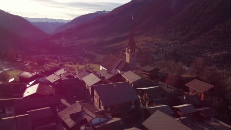 Flying-over-the-roofs-of-Ernen,-Switzerland-at-sunset--Beautiful-orbiting-flight-showing-the-old-church-and-mountain-valley