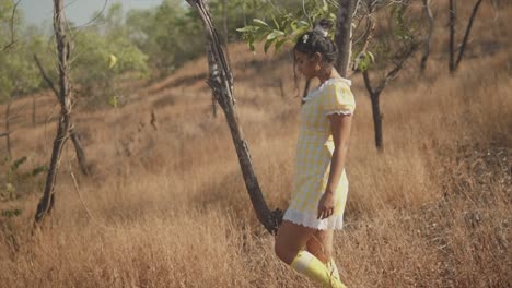 An-attractive-Asian-female-wearing-a-pretty-yellow-checked-dress-dancing-alone-carefree-around-a-tree-in-the-middle-of-a-remote-dry-grass-field,-India
