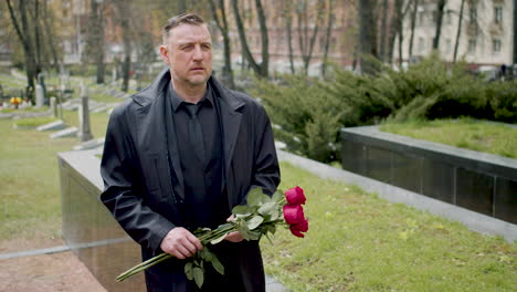 Caucasian-man-in-black-raincoat-and-suit-holding-roses-while-walking-in-a-graveyard