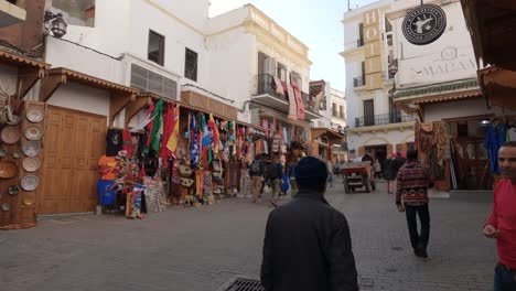 Einheimische-Und-Touristen-Gehen-über-Den-Kleinen-Platz-Von-Tanger,-Vorbei-An-Einem-Geschäft,-Das-Landesflaggen-Verkauft
