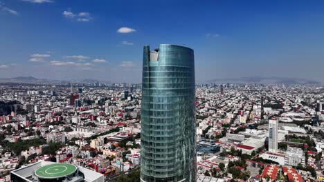 Aerial-view-rising-towards-the-Torre-Mitikah-tower,-revealing-a-helipad-on-the-top,-in-sunny-Mexico-city