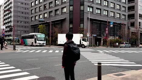 Joven-Asiático-Masculino-Con-Máscara-Y-Mochila-Esperando-En-El-Paso-De-Cebra,-Kyoto,-Japón