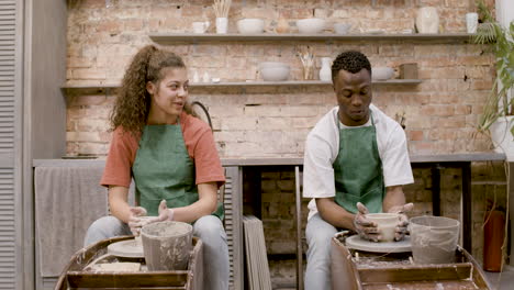 Front-view-of-employees-wearing-green-apron-modeling-ceramic-pieces-on-potter-wheel-in-a-workshop-while-talking-to-each-other