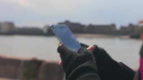 Close-up-shot-of-a-woman-walking-while-typing-on-her-smartphone-near-a-river-with-the-city-in-the-background