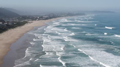 Südafrikas-Wildnisküste,-Langer-Sandstrand-Mit-Vielen-Meereswellen,-Die-An-Land-Krachen,-Und-Strandhäusern-Im-Hintergrund