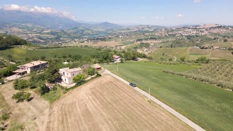 Wide-angle-drone-shot-tracking-a-vehicle-driving-in-the-remote-countryside-of-Italy-surrounded-by-a-beautiful-vista-of-vineyards,-olive-trees-and-a-mountain-range-located-in-Abruzzo,-Italy