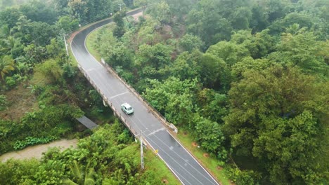 Sich-Schnell-Bewegende-Luftaufnahme-über-Dem-Kopf,-Die-Einem-Weißen-Auto-Auf-Einer-Nassen,-Windigen-Straße-Durch-Handaganawa-In-Sri-Lanka-Folgt