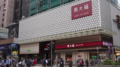 La-Gente-Camina-Por-La-Calle-En-Hong-Kong,-El-Logotipo-De-Chow-Tai-Fook-En-El-Edificio