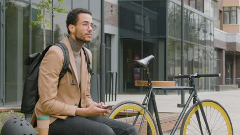 Afroamerikanischer-Mann-In-Formeller-Kleidung-Mit-Handy,-Während-Er-Neben-Seinem-Fahrrad-Auf-Einer-Holzbank-Auf-Der-Straße-Sitzt