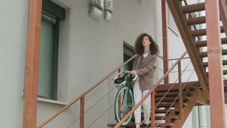 Lockige-Kaukasische-Frau-In-Formeller-Kleidung,-Die-Ihr-Fahrrad-Die-Treppe-Hinunter-Trägt