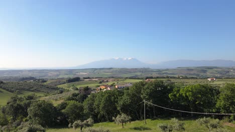 Weitwinkel-Drohnenaufnahme-über-Olivenbäume-Und-Ein-Kleines-Dorf,-Das-Sich-An-Einem-Frühlingsmorgen-In-Richtung-Des-Majella-Gebirges-In-Der-Landschaft-Der-Abruzzen-In-Italien-Bewegt