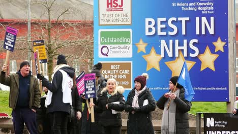 Huelga-De-Personal-Del-Nhs-Con-Exceso-De-Trabajo-En-La-Línea-De-Piquete-Ondeando-Pancartas-Y-Banderas-En-El-Hospital-En-St-Helens-En-Protesta-Exigiendo-Un-Salario-Justo