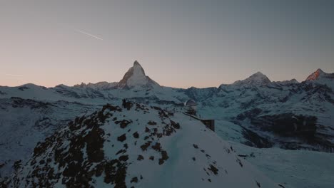 Sideways-Drone-shot-Over-Gornergrat-Summit-with-stunning-view-of-Matterhorn-Zermatt-in-Swiss-Alps-at-Sunset-during-winter-with-golden-clear-sky-and-airplane-flying-over