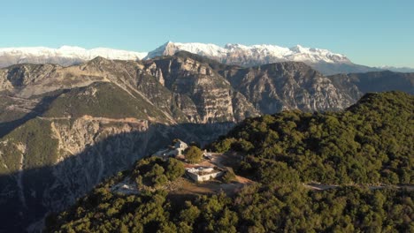 Drohnenvideo,-Das-über-Eine-Kirche-Auf-Einem-Berg-Fliegt,-Und-Die-Schneebedeckten-Gipfel-Des-Berges-Tzoumerka-In-Griechenland-In-Der-Ferne-Und-Ein-Tiefer,-Steiler-Canyon-Klippenschluchtfluss-Unten
