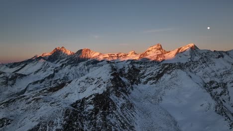 Sideways-Rotating-Aerial-Drone-Video-of-mountain-tops-during-sunset-with-full-moon-rising-and-stunning-colours-in-clear-sky-over-breathtaking-beautiful-swiss-alps