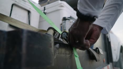 Ratcheting-down-coolers-with-green-tie-down-strap-onto-truck-trailer,-close-up-slow-motion-4k-tracking-shot-of-African-American-man's-hands-tightening