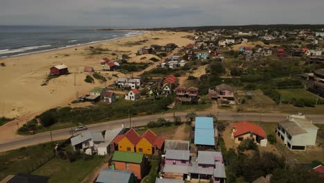 Aerial-flyover-colorful-houses-in-front-of-Atlantic-Oceana-and-sandy-beach-of-Punta-del-Diablo,-Uruguay