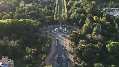 Carretera-Circular-Entre-El-Bosque-Verde-De-Mendoza,-Argentina,-Los-Autos-Conducen-Alrededor-Del-Cruce-De-La-Rotonda,-Las-Copas-De-Los-árboles-Y-La-Arquitectura-De-La-Ciudad-Del-Famoso-Destino-Del-Vino