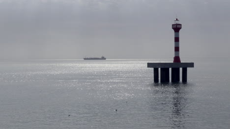 Lighthouse-in-the-fishing-port-of-Bandim,-with-a-large-boat-approaching-in-the-background-on-a-misty-morning