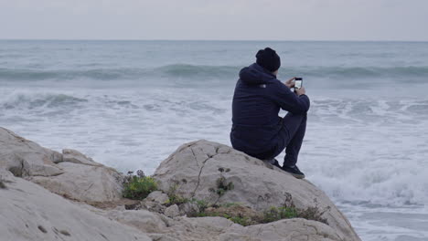 Man-sitting-on-a-rock-taking-photos-with-his-phone-of-stormy-sea-waves-crushing-in-front-of-him-before-departing-from-the-scene