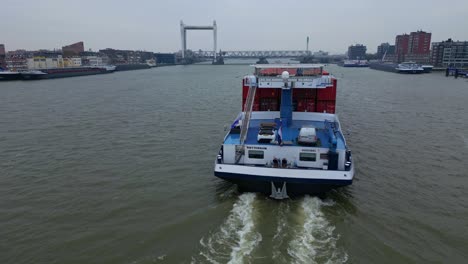 Aerial-View-Off-Stern-Of-Belicha-Inland-Freighter-Transporting-Intermodal-Containers-Along-Oude-Maas-With-View-Of-Spoorbrug-Railway-Bridge-In-background