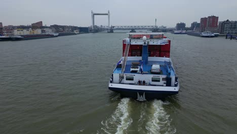 Luftaufnahme-Vom-Heck-Des-Binnenfrachters-Belicha,-Der-Intermodale-Container-Entlang-Der-Oude-Maas-Transportiert,-Mit-Blick-Auf-Die-Eisenbahnbrücke-Spoorbrug-Im-Hintergrund