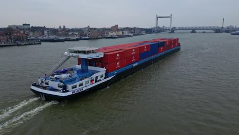Aerial-Shot-Of-Belicha-Inland-Freighter-Transporting-Intermodal-Containers-Along-Oude-Maas-With-View-Of-Spoorbrug-Railway-Bridge-In-background