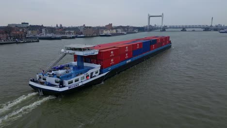 Toma-Aérea-Del-Carguero-Interior-Belicha-Que-Transporta-Contenedores-Intermodales-A-Lo-Largo-De-Oude-Maas-Con-Vista-Al-Puente-Ferroviario-Spoorbrug-En-Segundo-Plano