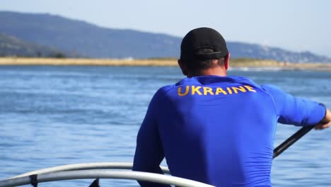 Slow-motion-shot-from-behind,-of-a-Paralympic-athlete-rowing-a-river-on-a-disabled-canoe,-wearing-a-blue-Ukraine-shirt