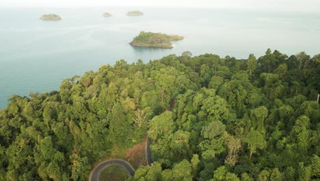 aerial-shot-of-rainforest-with-ocean-and-islands-in-the-distance,-Koh-Chang,-Thailand,-shot-at-sunrise-with-a-windy-road-in-shot